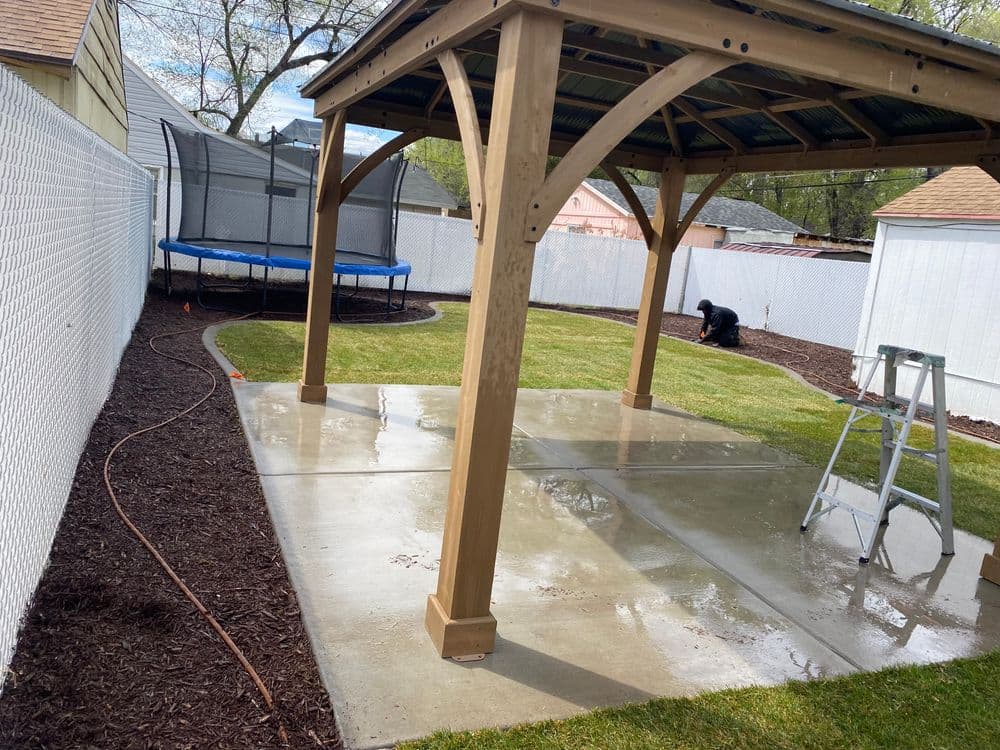 Backyard with patio pavilion, freshly cleaned concrete, trampoline, and landscape maintenance in progress.