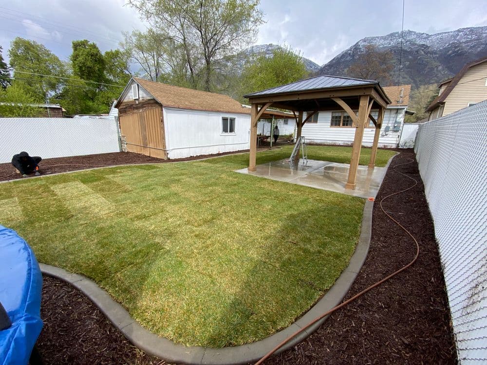 Newly landscaped backyard with green lawn, gazebo, and garden shed against a mountain backdrop.