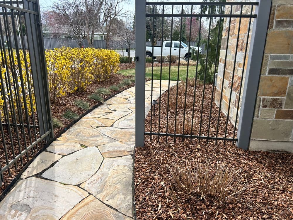 Stone path leading through a metal gate with landscaped bushes and mulch in a garden setting.