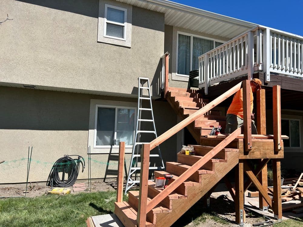 Wooden deck stairs under construction with a ladder and tools visible, bright blue sky.