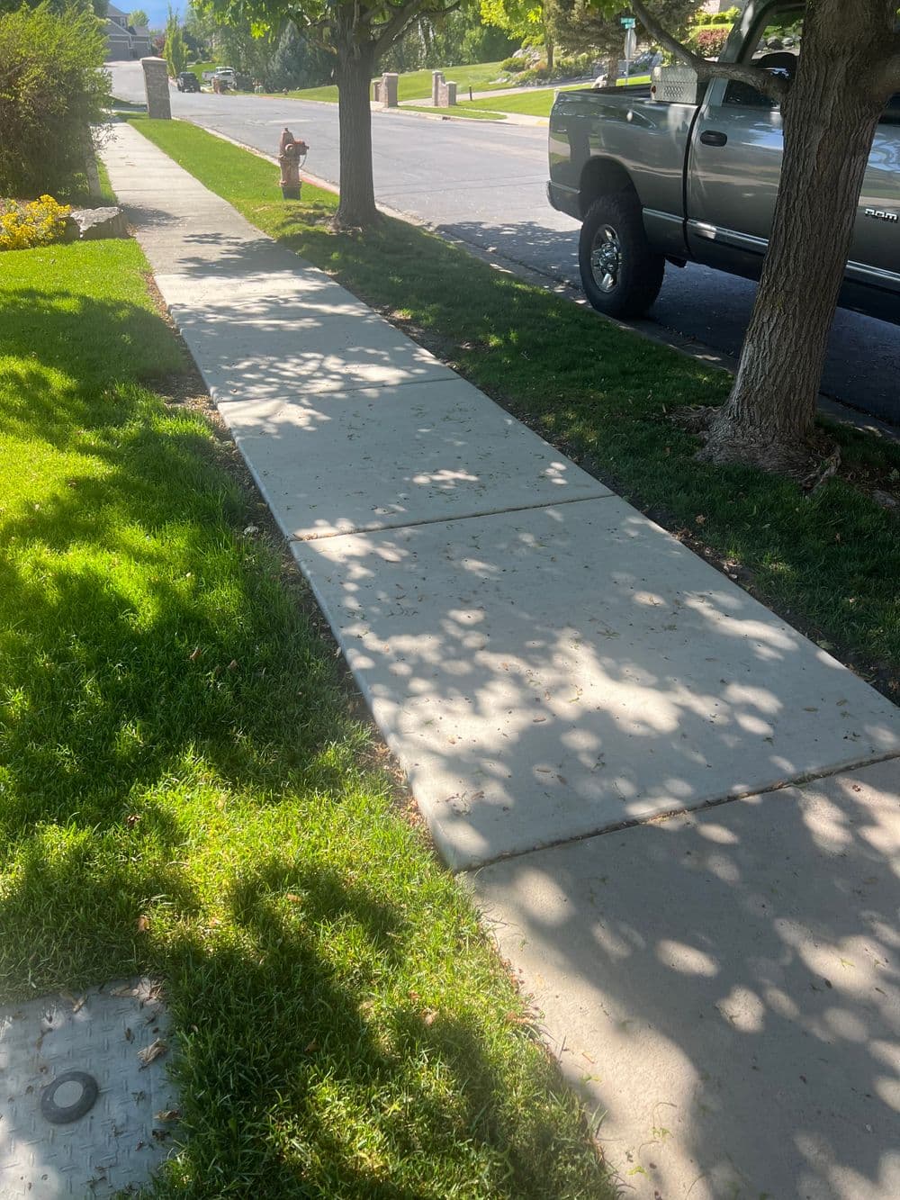 Concrete sidewalk bordered by grass and trees, with a truck parked nearby on a sunny day.
