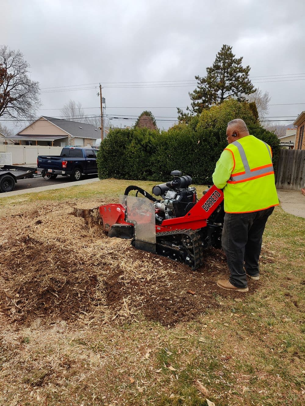 Man in safety vest operating a stump grinder on a residential lawn, cloudy skies above.