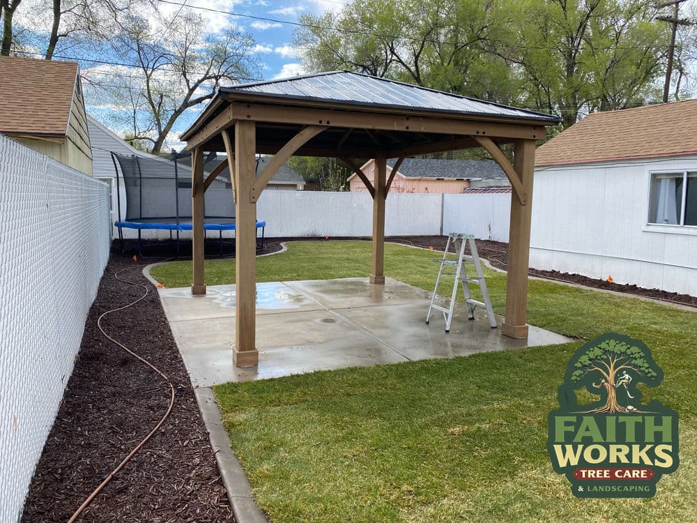 Wooden gazebo in a backyard with grass, trampoline, and ladder on a patio surface.